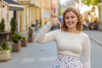 Fototapeta premium Like. Happy redhead woman raises thumbs up agrees with something or gives positive reply recommends advertisement likes good idea feedback celebrating success victory outdoors. Girl in city street.