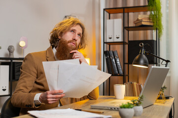 Caucasian redhead freelancer accountant working with financial graph documents, calculate taxes for charts and reports at home office desk. Business man guy working on laptop, planning expenses budget