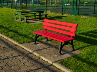 A red bench sits in a grassy area next to a playground fence. The bench is empty and the grass is green