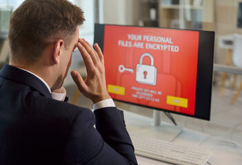 Stressed businessman faces ransomware on computer monitor. In an office, encryption and malware and virus alerts signal a cyberattack and extortion. Clear concept of cyber extortion and stress.