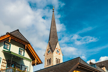 The slender, grey spire of the Hallstatt Lutheran Church rises elegantly against a bright blue sky, flanked by traditional alpine rooftops.