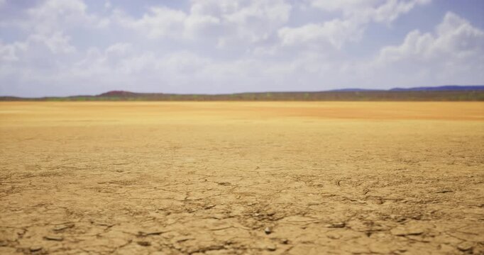 A vast dry landscape stretches out, with cracked earth and muted colors under a clear blue sky and fluffy clouds, creating a serene yet desolate atmosphere.