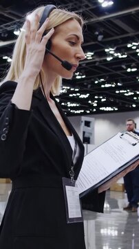 Event coordinator caucasian woman wearing headset clipboard monitors busy convention hall communicating via microphone while checking schedule, coordinating exhibitors and staff, adjusting lanyard