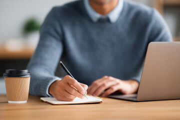 Young person writing notes in notebook with pen while working on laptop at wooden desk with coffee cup nearby