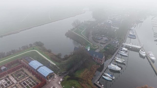 Aerial view of foggy riverside formal garden with greenhouses, red brick building with clock tower, canal, marina with docked boats, and quiet residential zone blend nature and architecture.