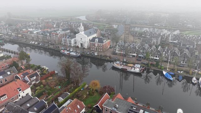 Aerial view of foggy Dutch canal town gabled brick homes, docked boats, and tall church tower form a tranquil, symmetrical waterfront scene softened by mist.