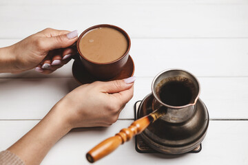 Close-up of female hands holding ceramic coffee cup next to Turkish cezve on white rustic table