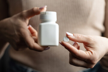 Close-up of female hands holding white pill bottle and single white round pill with score line, concept of medication and pharmaceutical treatment