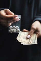 Close-up of female hands holding cash and transparent plastic bag with white narcotic pills, concept of drug abuse and drug trafficking
