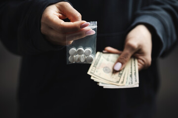 Close-up of female hands holding cash and transparent plastic bag with white narcotic pills, concept of drug abuse and drug trafficking