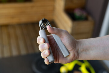 Close-up of a strong male hand squeezing a heavy-duty silver grip strengthener for armwrestling wrist training in a bright home gym. Background shows yellow bands, weight plates and fitness gear