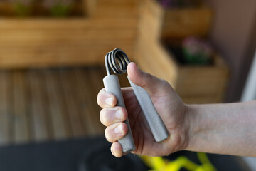 Close-up of a strong male hand squeezing a heavy-duty silver grip strengthener for armwrestling wrist training in a bright home gym. Background shows yellow bands, weight plates and fitness gear