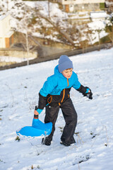 Active little boy in warm winter clothes holding a blue sled while walking uphill on a snowy slope, enjoying outdoor winter fun and play.