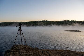  A camera on a tripod perched on a rocky cliff captures the tranquil early morning scene, under a clear sky with soft sunrise light. A serene lakeside landscape at dawn with mist rising over calm wate