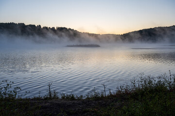 Golden morning mist rises from the calm waters of Lake Vlasina in Serbia, surrounding a small vegetated islet with a forested shoreline in the background.