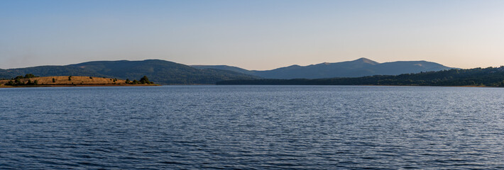 A minimalist alpine landscape depicting a tranquil lake beneath bluish mountain ranges. The soft light, clear horizon, and natural color palette evoke associations with untouched wilderness.