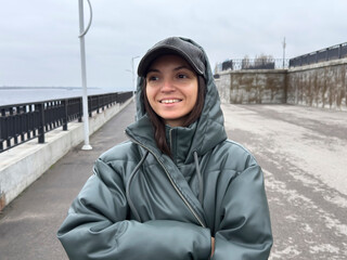 A woman stands on a path by the water, smiling and looking ahead. The sky is cloudy