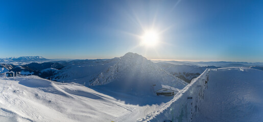 Wide panoramic view of snow-covered peaks in the Low Tatras National Park, Slovakia, featuring a bright sun star, frozen mountain ridges, and a distant view of the High Tatras under a clear winter sky