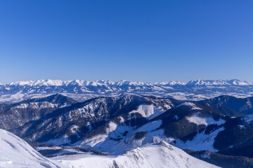 Wide panoramic view of snow-covered peaks in the Low Tatras National Park, Slovakia, featuring a bright sun star, frozen mountain ridges, and a distant view of the High Tatras under a clear winter sky