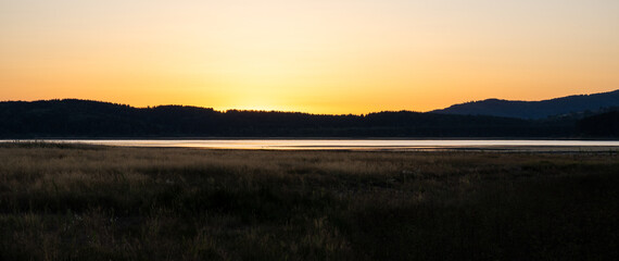 Minimalist dusk landscape featuring tall dry meadow grass, a reflective lake, and distant forested hills. 