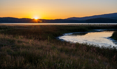 Golden sunset over a calm lake with wide open grassland in the foreground and dark rolling hills on the horizon. The clear sky fades from warm orange to soft blue.