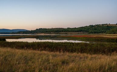 Minimalist dusk landscape featuring tall dry meadow grass, a reflective lake, and distant forested hills. 