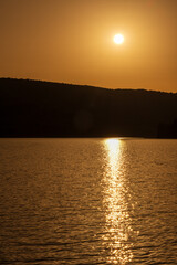 A golden sunset over calm lake waters with a reflective trail of light leading to the silhouettes of hills. Warm orange tones fill the sky, creating a serene twilight landscape ideal for sunset themes