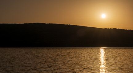 Golden hour over a quiet lake with the sun shining on reflective water and silhouetted hills on the horizon. Evocative sunset landscape with warm light and calm mood, suited for backgrounds.