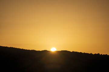 A golden sunset over calm lake waters with a reflective trail of light leading to the silhouettes of hills. Warm orange tones fill the sky, creating a serene twilight landscape ideal for sunset themes