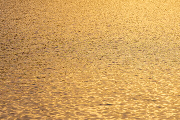 Golden sunlight reflects across gently rippling water, creating a shimmering abstract texture. Warm tones and soft wave patterns form a calming natural background.