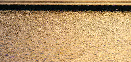 Golden sunlight reflects across gently rippling water, creating a shimmering abstract texture. Warm tones and soft wave patterns form a calming natural background.