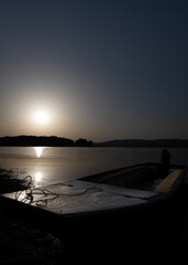 A landscape photo of a calm lake at dawn with gentle sun rays reflecting off the calm water and a fishing boat resting on the shore waiting for fishermen to set out to sea.