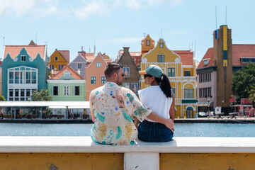 Under the sunny Caribbean sky, a couple shares a joyful moment by the colorful waterfront of Willemstad, Curacao. The charming pastel buildings create a picturesque backdrop, perfect for romance.