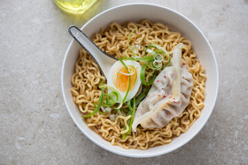 Bowl of ramen noodles with boiled egg and steamed dumpling on a beige stone background, horizontal shot, high angle view