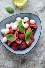 Bowl of strawberry caprese with mini mozzarella and green basil, vertical shot on a light-brown granite background