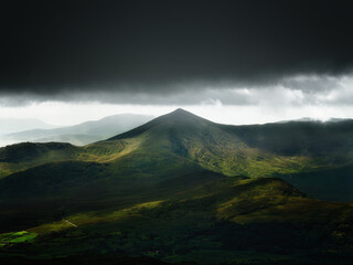 A stunning view of rugged mountain peaks in Ireland. Dark storm clouds loom above, while patches of sunlight illuminate the green valleys.