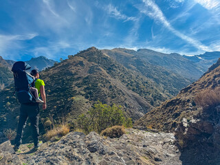 Fototapeta premium Male hiker carrying a toddler in a backpack stands on a rocky ridge admiring the majestic mountain peaks of Sierra Nevada in Andalusia, Spain, during sunny trekking adventure on Verada de la Estrella.
