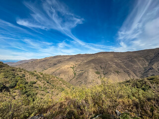 The rugged brown slopes of the Sierra Nevada mountains rise beneath a deep blue sky with dramatic clouds, contrasting with the green valley vegetation along the Vereda de la Estrella in Spain.