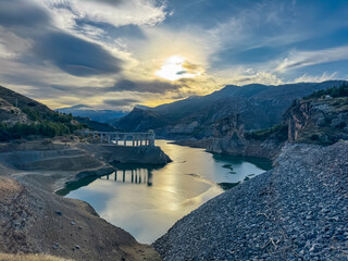 A view of the rocky banks of Canales Reservoir leading to calm waters under a bright sunburst, capturing the dramatic mountain landscape of the Sierra Nevada near Granada in Andalusia, Spain.