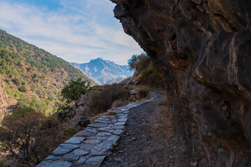 A stone path winds beneath a dark rocky overhang along the steep slopes of the Genil valley, offering views of the Sierra Nevada peaks on the Vereda de la Estrella trail in Granada, Andalusia, Spain.