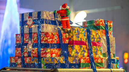 Small Santa Claus figure attached to colorful gift boxes with festive lights