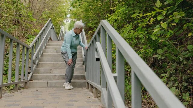 Elderly woman standing on outdoor stairs and holding her knee due to sudden pain