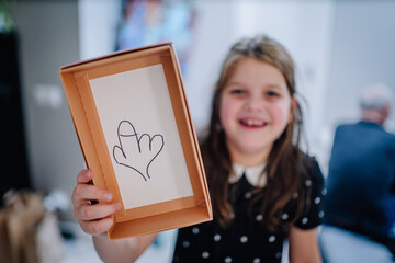 Young girl with brown hair, wearing a polka dot dress, joyfully holds a box displaying a playful drawing, capturing a moment of creativity and fun in a bright indoor setting