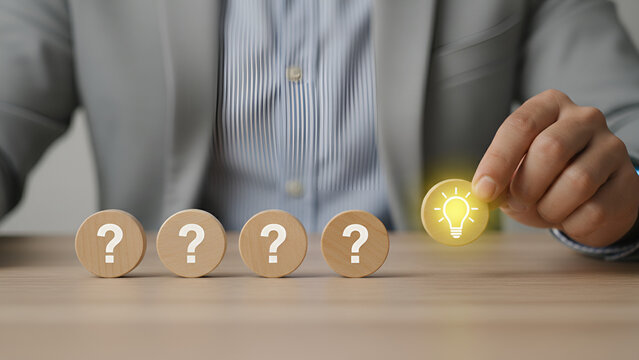 Businessman holds a glowing lightbulb icon while several question mark blocks rest on the wooden desk representing problem solving