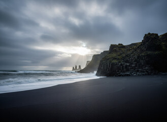 Black sand beach with basalt columnar cliff and sea stack formation under dramatic cloudy sky with sun rays shining through