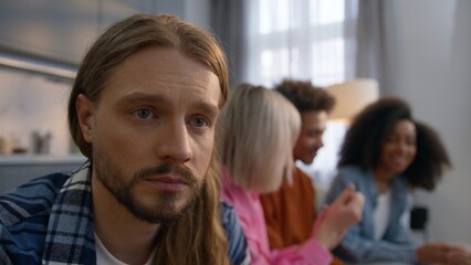 Obraz premium Stressed sad Caucasian man sitting couch home thinking about problems depressed male feeling separated ignoring friends while multiracial African women men talk together friendship loneliness anxiety