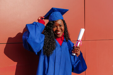 Black graduate student smiling while holding her diploma and adjusting her academic cap