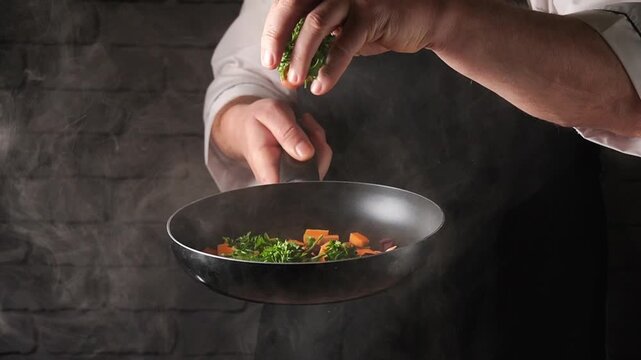 A chef sprinkles fresh herbs or parsley into a skillet with colorful vegetables while cooking in a kitchen. Steam rises from the pan as the chef focuses on the dish.