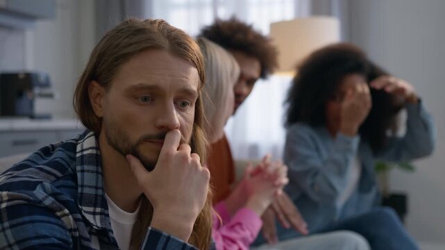 Sad anxious Caucasian man deep thoughts at home male isolated ignoring friends while multiethnic African women men talking together loneliness overthinking mental struggle indoors blurred background