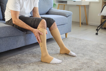 Man using compression stockings for varicose veins therapy at home. Person sitting on sofa,...